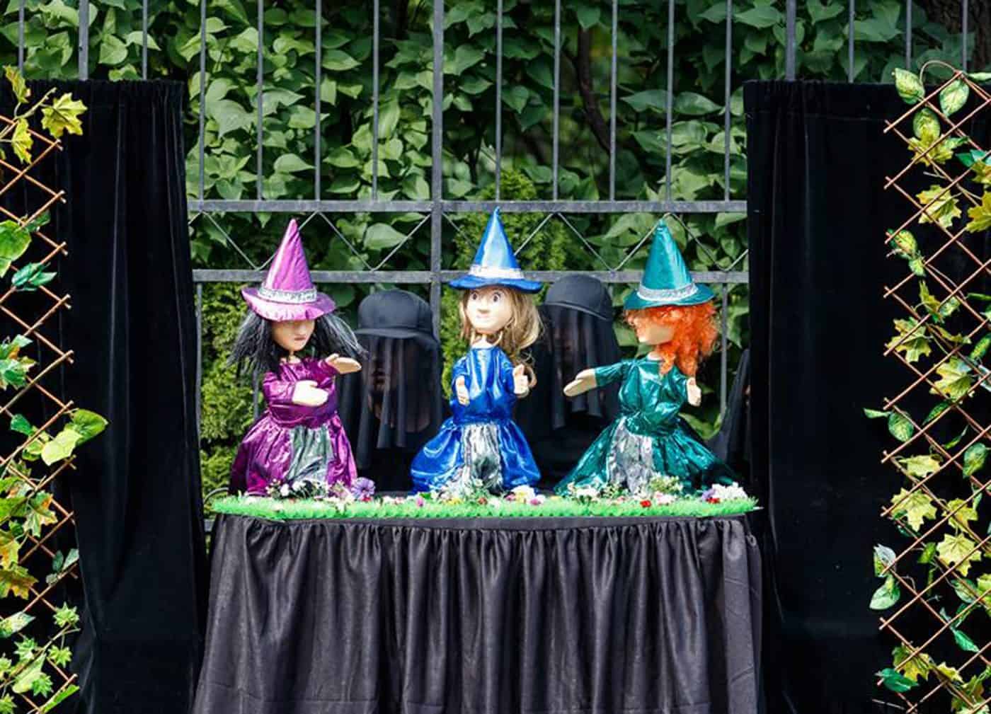Magicienzi de păpuși cu pălării colorate și costume strălucitoare, decor de grădină.
