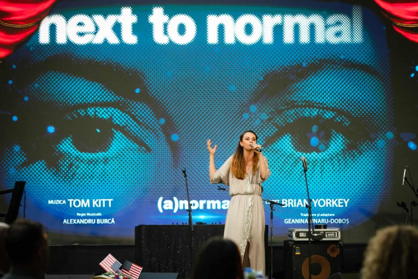 Next to normal, spectacol muzical pe scenă cu o solistă în față, microfon și public în prim-plan.