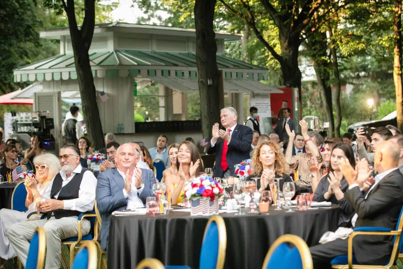 Eveniment în aer liber cu oameni eleganți și discursuri, în parc cu copaci și lumină naturală.
